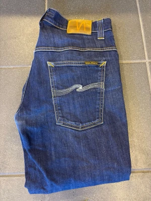 Nudie Jeans- W36 - Mörkblå - Säljer ett par snygga mörkblå jeans från Nudie Jeans. Smal passform med stretch som sitter bekvämt och följer kroppen fint. Tvättad ljus denim med naturligt slitna detaljer och klassiska Nudie-sömmar på bakfickorna. Jeansen är i fint använt skick utan större anmärkningar. Perfekta för vardagsbruk och enkla att matcha med t-shirt, skjorta eller hoodie. ✅ Märke: Nudie Jeans ✅ Färg: Mörkblå denim ✅ Passform: Slim fit / smal modell ✅ Skick: Bra begagnat skick ✅ Stretchig och bekväm kvalité 