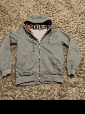 Grå zip-hoodie från Burberry - Snygg grå hoodie från Burberry med dragkedja och klassiskt rutigt foder i huvan. Mjukt material, två fickor fram och ribbade muddar. Perfekt för en chill och stilren look. Ikonisk Burberry-detalj i huvan ger extra lyxkänsla.