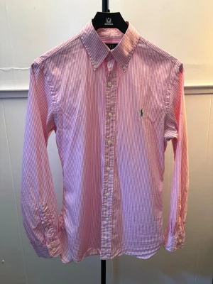 Rosa och vit randig skjorta Polo Ralph Lauren - Klassisk långärmad skjorta från Polo Ralph Lauren i rosa och vitt randigt mönster. Skjortan har knappar framtill, button-down krage och den ikoniska gröna logotypen broderad på bröstet. Tillverkad i bomull för en skön och luftig känsla.