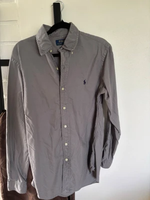 Grå skjorta från Polo Ralph Lauren - Klassisk grå skjorta från Polo Ralph Lauren med broderad logga på bröstet. Skjortan har knappar framtill, button-down krage och långa ärmar. Tillverkad i mjuk bomull för en bekväm känsla och stilren look.