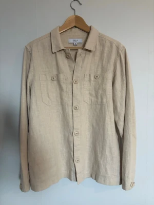 Beige linneskjorta overshirt från ERLA - Stilren beige overshirt från ERLA i luftigt linne. Skjortan har två bröstfickor med knapp, klassisk krage och knäppning framtill. Perfekt lager på lager-plagg med avslappnad passform och långa ärmar.
