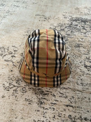 Burberry bucket hat i klassiskt rutmönster - Säljer en snygg bucket hat från Burberry i beige med det ikoniska rutmönstret i svart, vitt och rött. Hatten är tillverkad i bomull och har en avslappnad passform. 