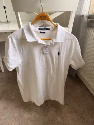 Vit pikétröja från Polo Ralph Lauren - Klassisk vit pikétröja från Polo Ralph Lauren med kort ärm och krage. Tröjan har en broderad logga på bröstet och är tillverkad i mjuk bomull. Perfekt för en clean och stilren look. Skulle säga att den sitter som L/M