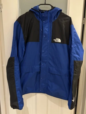Blå vindjacka från The North Face - Snygg blå vindjacka från The North Face med svarta detaljer på axlar och ärmar. Jackan har huva, dragkedja och tryckknappar framtill samt två stora fickor. Tillverkad i lätt och slitstarkt material, perfekt för blåsiga dagar.