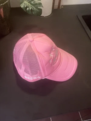 Rosa truckerkeps med mesh och brodyr - Säljer en rosa truckerkeps från Capster med meshpaneler och böjd skärm. Kepsen har vit brodyr fram och bak samt justerbar passform. Perfekt för dig som vill ha en sportig och trendig look.
