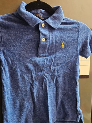 Blå pikétröja från Polo Ralph Lauren till barn - Klassisk blå pikétröja från Polo Ralph Lauren med gul broderad logga på bröstet. Tröjan har korta ärmar, krage och två knappar framtill. Perfekt till en avslappnad men ändå stilren look.