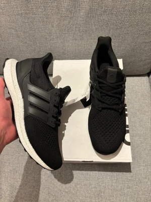 Adidas Ultraboost 1.0 Svarta Sneakers - Säljer ett par svarta Adidas Ultraboost 1.0 sneakers med vit sula och klassisk tre-randig design på sidan. Ovandelen är i ventilerande mesh och skon har snörning för perfekt passform. Perfekta för dig som vill ha både stil och komfort.