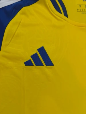 Svensk landslagströja Adidas gul/blå - Gul svensk landslagströja från Adidas med blå detaljer och broderad landslagssköld på bröstet. Tröjan har blå Adidas-logga, rund halsringning och vita ränder på axlarna. Tillverkad i lätt funktionsmaterial, perfekt för fotboll eller träning.