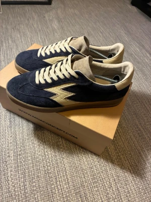 Mörkblå sneakers från MDA Concept - Snygga mörkblå sneakers från Moa Concept med beige detaljer och blixtlogga på sidan. Skorna har beige snörning, beige hälparti och plös i mocka. Yttermaterialet är mocka och sulan är i gummi. Perfekta för dig som gillar retrovibbar och unik design. Har använts fåtal gånger då jag köpte dem från NK 2026-05-08. Ser helt nya ut men texten på ena skon har lossnat lite som ni ser på en av bilderna. Nypris 2800. Tveka inte kring frågor.