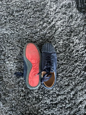 Blåa Louboutin sneakers med nitar - Snygga blåa sneakers från Christian Louboutin i mocka med ikoniska röda sulor och coola nitar på tån. Skorna har blå snören och lågt skaft, perfekt för dig som vill sticka ut med din stil.
