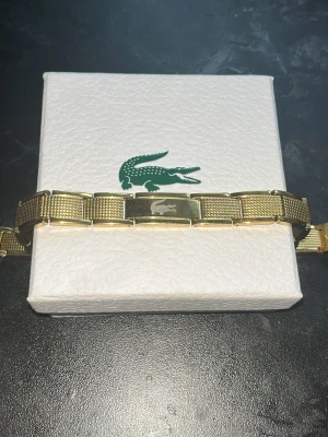 Lacoste Armband Guldfärgat  - Snyggt Lacoste guldfärgat armband med länkdesign. Armbandet har en stilren och modern look med matta och blanka partier i metallen. Perfekt accessoar för dig som gillar klassisk men trendig stil.  Fick den i present och har inte kvitto för jag har inte frågat om kvittot för att föräldrar vet inte att jag säljer den. 