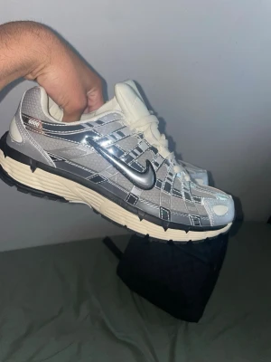 Nike P 6000 silver sneakers - Skorna är äkta, skanna QR koden.                   Snygga Nike P6000 sneakers i silver och grå med vita detaljer och svart sula. Skorna har mesh och syntetmaterial i ovandelen, samt reflekterande detaljer och klassisk Nike Swoosh på sidan. Perfekta för dig som gillar retrostil och streetwear.