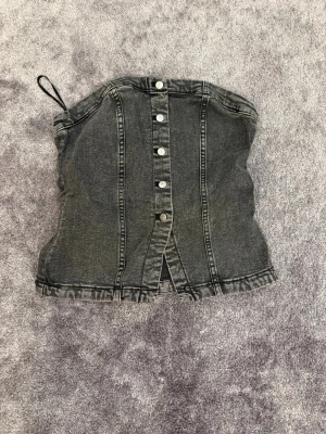 Svart denim tubtopp med knappar - Cool svart tubtopp i denim med knappar framtill och en liten slits nedtill. Toppen har en bandeauformad passning och markerade sömmar som ger en edgy look. Perfekt att styla med höga jeans eller kjol för en trendig outfit.