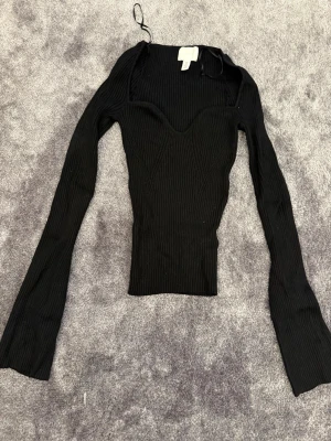 Svart ribbad långärmad topp - Trendig svart ribbad topp med lång ärm och markerad v-ringning. Figurnära passform och stretchigt material som ger en snygg siluett. Perfekt att styla med jeans eller kjol för en cool look.