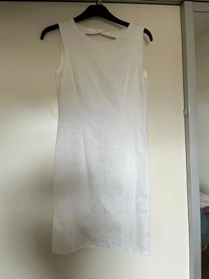 Vit ärmlös kortklänning med mönster - Säljer en elegant vit kortklänning med diskret mönster och rund halsringning. Klänningen är ärmlös och har en snygg dragkedja i ryggen samt en öppen detalj vid nacken. Perfekt för sommarens alla tillfällen såsom midsommar eller studenten/examen använd 1 gång och skräddarsydd för att passa en S