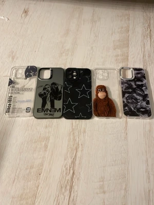 Fem unika mobilskal i olika stilar - Säljer fem coola mobilskal till iPhone 12 Pro Max: ett genomskinligt med Red Bull Coconut Edition-tryck, ett svart Eminem-skal, ett svart skal med vita stjärnor, ett genomskinligt skal med brun fluffig apa och ett genomskinligt skal med svartvitt kamouflagemönster. Perfekt för dig som vill variera stilen på mobilen.