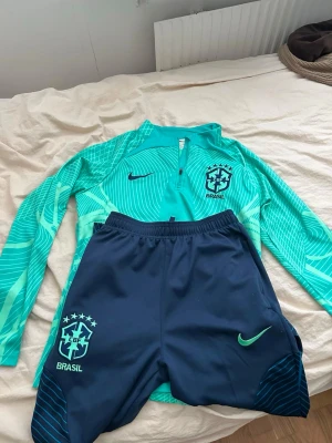 Nike Brasil träningsset långärmad - Snyggt träningsset från Nike med Brasil-logga. Tröjan är turkos med grafiskt mönster, lång ärm och halv dragkedja. Byxorna är mörkblå med turkosa detaljer och elastisk midja. Perfekt för fotbollsträning eller gymmet.