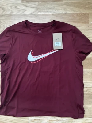 Vinröd Nike t-shirt med logga - Snygg vinröd t-shirt från Nike med klassisk stor logga i vitt och rött på bröstet. T-shirten har rund halsringning och korta ärmar. Perfekt för en sportig och avslappnad stil.