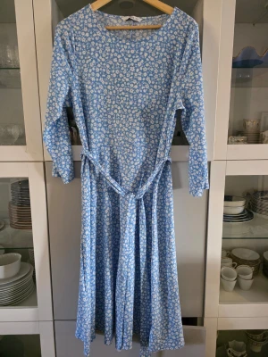 Blommig blå långklänning med knytband - Söt blå långklänning med vitt blommönster och knytband i midjan. Klänningen har lång ärm och rund halsringning. Perfekt för vår och sommar med sitt lätta och luftiga tyg. Ingen synlig logga eller märke. Från Ellos 