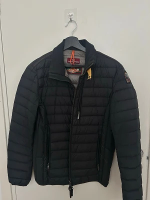 Svart pufferjacka från Parajumpers - Säljer en svart pufferjacka från Parajumpers i storlek S. Jackan är superlätt, har hög krage, dragkedja framtill och två sidofickor med dragkedja. Klassisk patch på ärmen och snygga detaljer som orange ögla vid halsen. Perfekt för kalla vinterdagar.