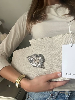 Beige kuvertväska med silverdetalj - Stilren beige kuvertväska i canvas med unik silverfärgad metallapplikation i form av ett löv på framsidan. Väskan har en asymmetrisk klaff och minimalistisk design som gör den till ett statement piece. Perfekt för dig som gillar trendiga accessoarer.