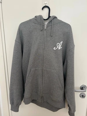 Grå zip hoodie från Axel Arigato - Snygg grå hoodie med dragkedja från Axel Arigato. Hoodien har en vit broderad A-logga på bröstet, justerbar huva med snören och stora fickor framtill. Tillverkad i mjuk bomullsblandning för extra komfort.