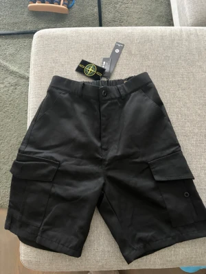Svarta cargoshorts från Stone Island - Snygga svarta cargoshorts från Stone Island med klassiska cargofickor på sidorna och knappstängning i midjan. Shortsen har en avslappnad passform och är tillverkade i bomull. Perfekta för sommaren och streetwear-stilen.