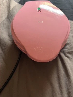 Rosa cake pop-maskin från C3 - Säljer en rosa cake pop-maskin från C3 med plats för 12 cake pops åt gången. Maskinen har en sladd och indikatorlampor på ovansidan. Perfekt för att baka små kakor eller bollar snabbt och enkelt hemma.