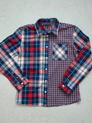 Rutig skjorta från Tommy Hilfiger stl 152 barn - Färgstark rutig skjorta från Tommy Hilfiger i blått, rött och vitt. Skjortan har lång ärm, klassisk krage och en bröstficka med kontrasterande rutmönster. Tillverkad i mjuk bomull och har vita knappar framtill. Obs barn storlek 152.