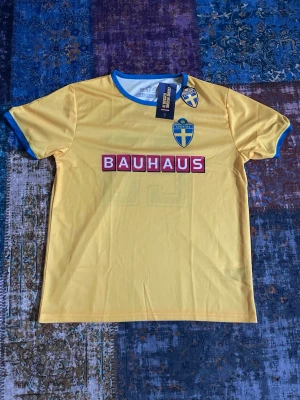 Svensk gul matchtröja euro 2020 - Gul matchtröja inspirerad av svenska landslaget med blå detaljer vid ärmslut och krage. Tryck med Bauhaus-logga och svensk flagga på bröstet. Tillverkad i lätt och ventilerande material, perfekt för sport och träning.