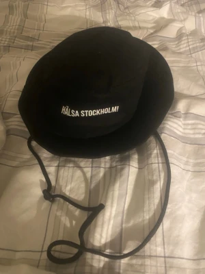 Svart bucket hat Hälsa Stockholm - Säljer en svart bucket hat från Sport Design Sweden med texten 'Hälsa Stockholm!' broderad framtill. Hatten har justerbar hakrem och är tillverkad i ett mjukt tyg som känns bekvämt på huvudet. Perfekt för dig som vill ha en stilren och enkel look.
