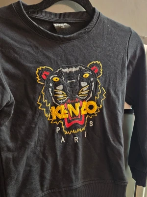 Svart Kenzo sweatshirt med tiger - Cool svart sweatshirt från Kenzo med det ikoniska tigerhuvudet broderat i gult, rött och vitt på bröstet. Tröjan har rund halsringning och ribbade muddar vid ärmslut och nederkant. Perfekt statement-plagg för dig som gillar streetwear.