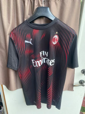 AC Milan matchtröja Puma XL - Snygg svart AC Milan matchtröja från Puma med röda och grå grafiska detaljer. Tröjan har klubbmärke, Puma-logga och Fly Emirates-tryck på bröstet. Tillverkad i lätt och ventilerande funktionsmaterial, perfekt för fotboll eller träning. Skänkt från Zlatan och använd på San Siro av honom!
