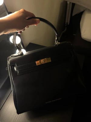 Svart handväska Victoria Hyde London - Stilren svart handväska från Victoria Hyde London i slätt konstläder. Väskan har ett elegant handtag, guldfärgad vridknapp och en klassisk fyrkantig form. Perfekt accessoar för dig som gillar en chic och tidlös look.