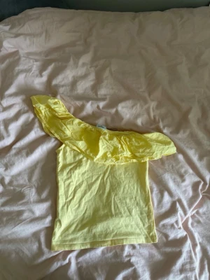 Gul oneshoulder topp med volang - Snygg gul oneshoulder topp med bred volangdetalj över axeln. Toppen är ärmlös och har en fräsch, ljus färg som passar perfekt till sommaren. Tillverkad i mjukt bomullsmaterial för en bekväm känsla.
