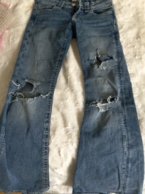 Blå slitna bootcut jeans - Säljer ett par ljusblå bootcut jeans med stora slitningar och hål på båda benen. Jeansen har klassisk femficksmodell och låg midja. Perfekta för en avslappnad och trendig look. Materialet är denim i bomull.