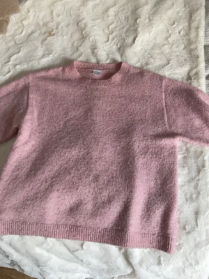 Ljusrosa stickad tröja - Mysig ljusrosa stickad tröja med rund hals och ribbade muddar. Tröjan har en enkel och klassisk design, perfekt för att hålla sig varm. Materialet ser ut att vara mjukt och fluffigt, vilket ger en skön känsla. Passar dig som gillar en avslappnad och stilren look.