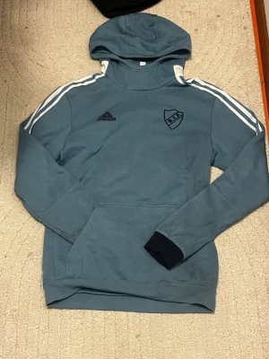 Blå Adidas hoodie med DIF-logga - Snygg blå hoodie från Adidas med Djurgårdens IF-logga på bröstet. Klassiska vita ränder längs ärmarna, huva och magficka. Mörkblå muddar och detaljer på ärmarna. Perfekt för dig som gillar sportig stil och vill representera DIF.
