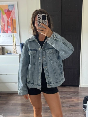 Oversized ljusblå jeansjacka - Säljer en oversized ljusblå jeansjacka med klassisk krage, två bröstfickor med lock och knappar framtill. Jackan har en avslappnad passform och raka ärmar. Perfekt att slänga över en hoodie eller t-shirt för en cool och avslappnad look.