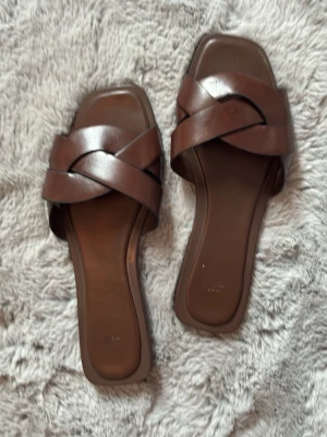 Bruna slip-in sandaler från H&M - Snygga bruna sandaler från H&M med breda korslagda remmar i konstläder. Modellen har fyrkantig tå och låg sula, perfekt för en avslappnad och trendig look. Passar till jeans, kjol eller shorts för en enkel och stilren outfit.