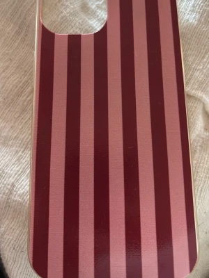 Randigt mobilskal iPhone 15 - Snyggt mobilskal med vertikala ränder i vinrött och rosa. Skalets form är klassisk med rundade hörn och ett stort kamerauttag. Tillverkat i hårdplast som ger bra skydd och stilren look till din mobil.