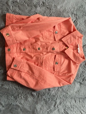 Korallfärgad jeansjacka från Cleo - Snygg korallfärgad jeansjacka från Cleo med klassisk krage, bröstfickor med lock och silverfärgade knappar. Jackan har raka sömmar och en fräsch, ungdomlig look som passar perfekt till vår och sommar.
