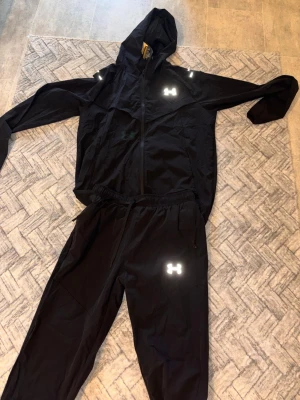 Svart träningsset från Under Armour - Snyggt svart träningsset från Under Armour med reflekterande loggor på både jacka och byxor. Byxorna passar i M 