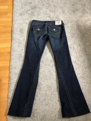 True Religion mörkblå bootcut jeans - Säljer ett par mörkblå jeans från True Religion med bootcut-modell. Jeansen har snygga kontrastsömmar, ficklock med knapp bak och klassisk True Religion-lapp i midjan. Tillverkade i denim med coola detaljer och retrovibbar.