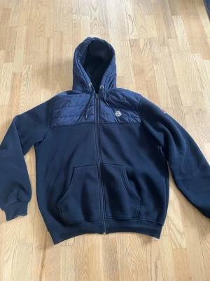 Svart hoodie från Moncler med dragkedja - Snygg svart hoodie/jacka från Moncler med dragkedja framtill och quiltad detalj över bröstet. Klassisk huva med dragsko och två stora fickor framtill. Moncler-logga på bröstet och ribbade muddar vid ärmar och nederkant. Perfekt för en chill och stilren look. Står på jackan att det är 3xl men jag skulle säga att det mer är xl. Använd en gång. Man kommer in på appen när man lägger telefonen mot loggan.
