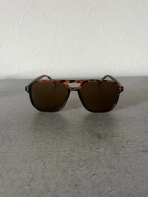 Solglasögon  - Snygga solglasögon med brunt leopardmönstrat båge och mörka glas. Klassisk aviator-inspirerad form som ger en cool vibe. Perfekta för soliga dagar och en trendig look. Tillverkade i syntetmaterial.