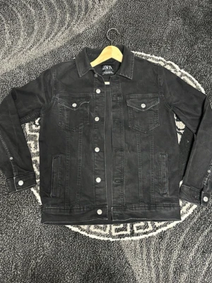 Svart jeansjacka från Zara - Stilren svart jeansjacka från Zara med klassisk krage, silverfärgade knappar och två bröstfickor med lock. Jackan har raka ärmar och diskreta sidofickor. Perfekt för en trendig och avslappnad look.