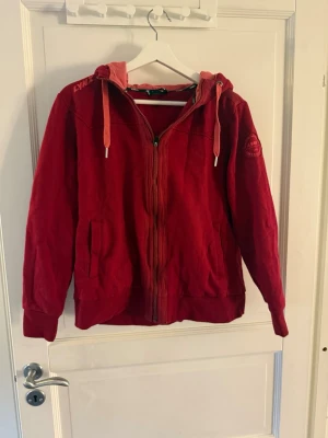 Röd hoodie med dragkedja och huva - Snygg röd hoodie från Lynx med dragkedja framtill, två fickor och justerbar huva med snören. Mjuk insida och ribbade muddar vid ärmslut och nederkant. Märkeslogga på ärmen 