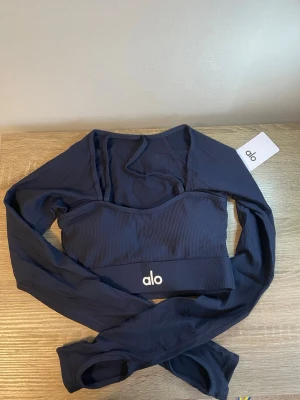 Mörkblå långärmad crop top från Alo - Snygg mörkblå långärmad crop top från Alo med ribbad struktur och logga framtill. Perfekt för träning eller yoga, med tight passform och stretchigt material som ger bra rörelsefrihet. Stilren design och bekväm känsla.