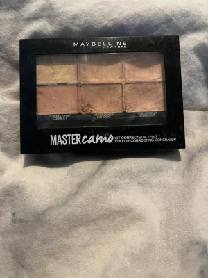 Maybelline Master Camo Concealer Kit - Palett med sex olika concealer- och korrigeringsnyanser från Maybelline. Innehåller både ljusa och mörka toner samt en highlighter, perfekt för att täcka ojämnheter och ge lyster. Kompakt svart plastförpackning med genomskinligt lock.